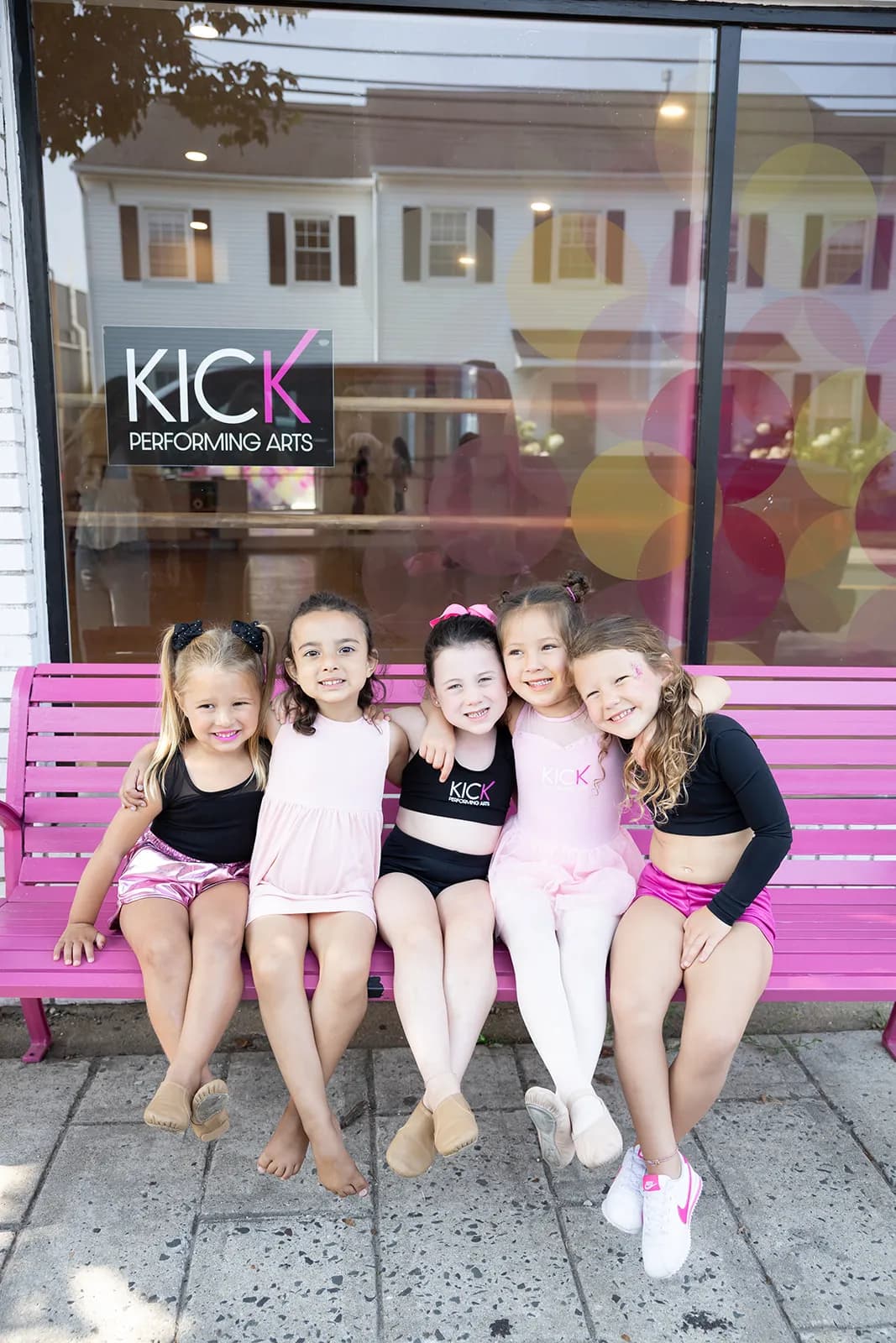 Young KICK dancers sitting together on a pink bench outside the studio
